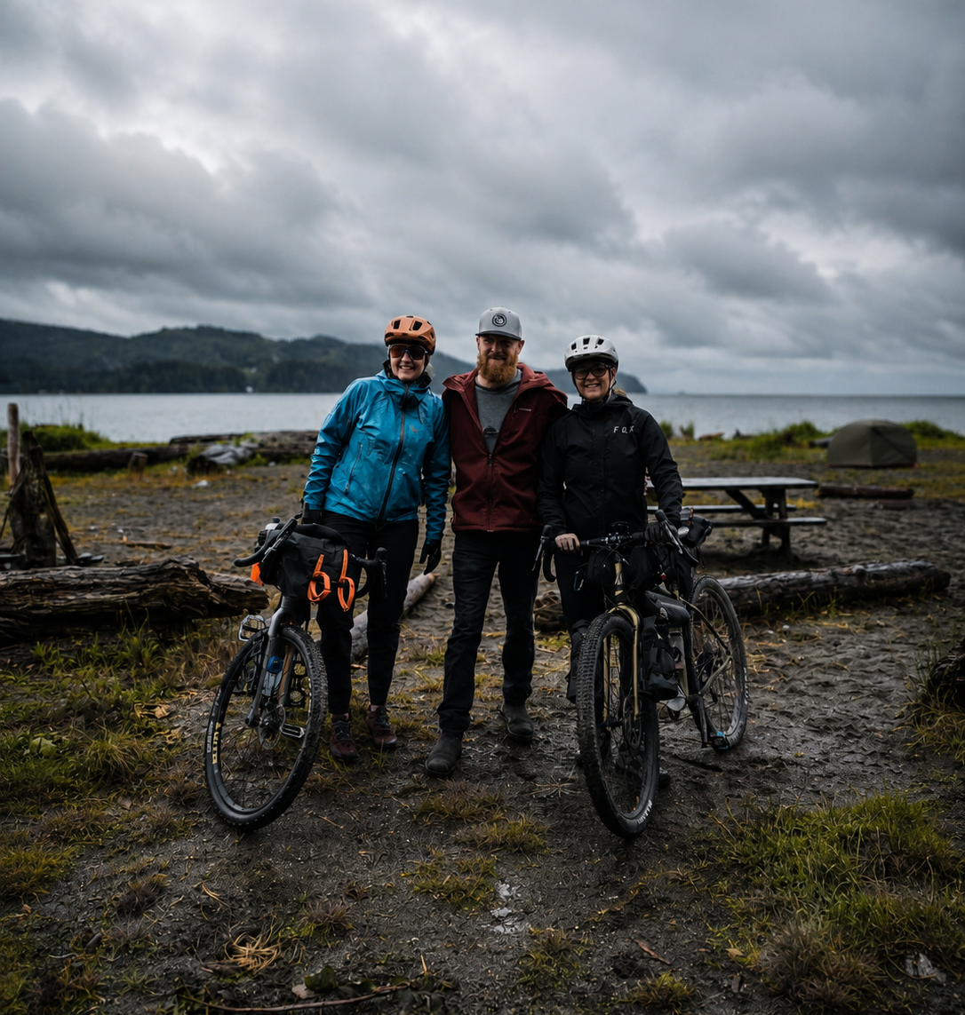 Three mountain bikers standing on a muddy lakeside trail under a cloudy sky, with two of them wearing helmets and standing next to their bikes.